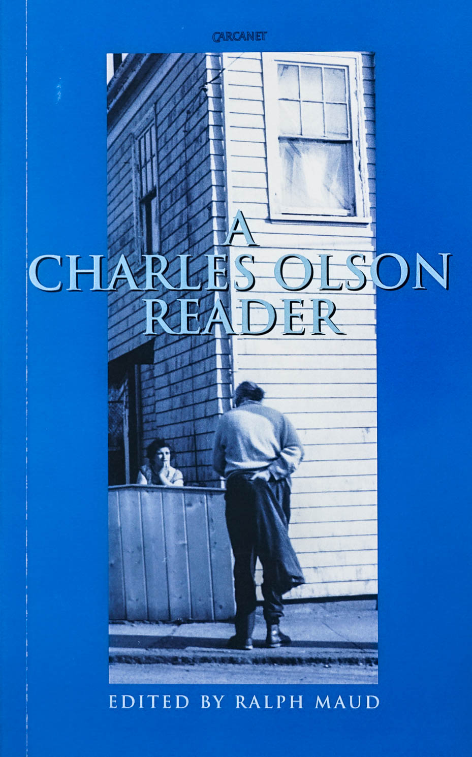 A Charles Olson Reader Editied by Ralph Maud in light blue serif type with an image of a man speaking to a women behind a fence and house and blue backdrop