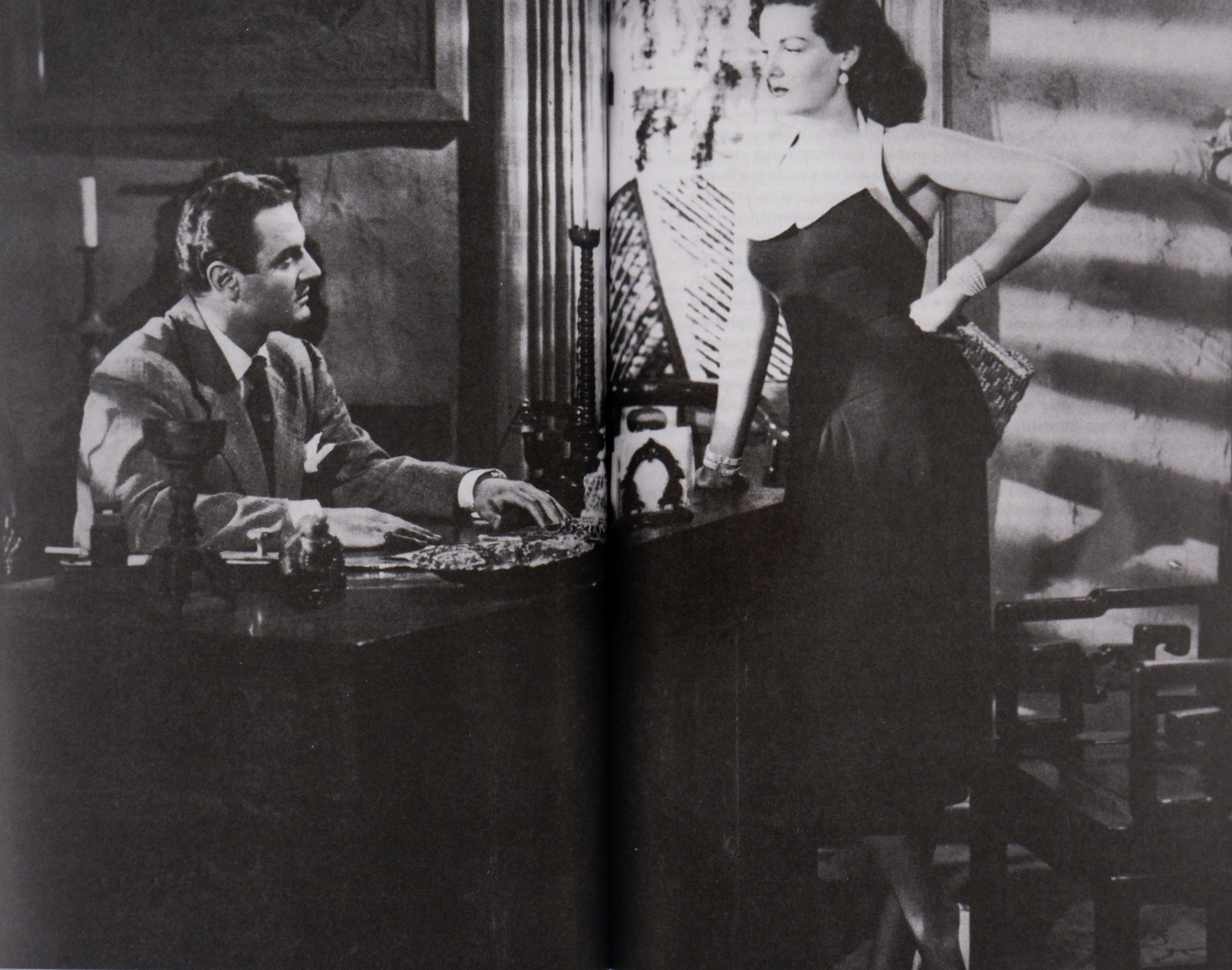 Spread of a movie still of a black-and-white film, a woman in an evening dress standing at a desk, looking down at a man in a suit.
