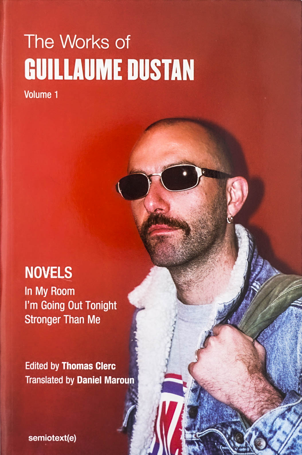 Image of the author standing in front of a red wall with aThe Works of Guillaume Dustan, Volume 1: In My Room; I'm Going Out Tonight; Stronger Than Me in white sans serif type 