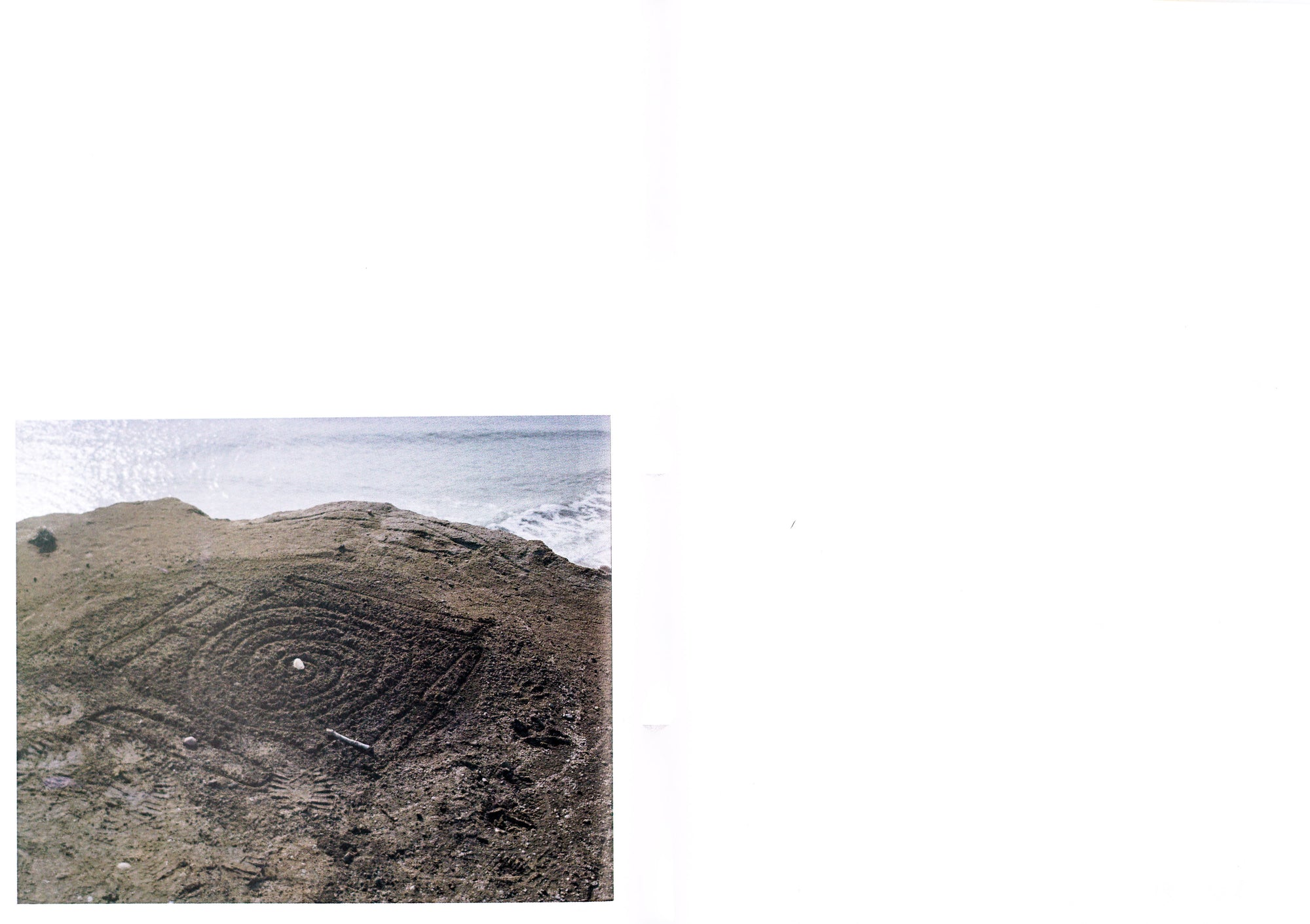 Book spread with white background, the right page is blank, on lower left corner of the left page is a colour photography of a coastal line with the sea in the background and a spiral pattern carved into the earth on the shore.